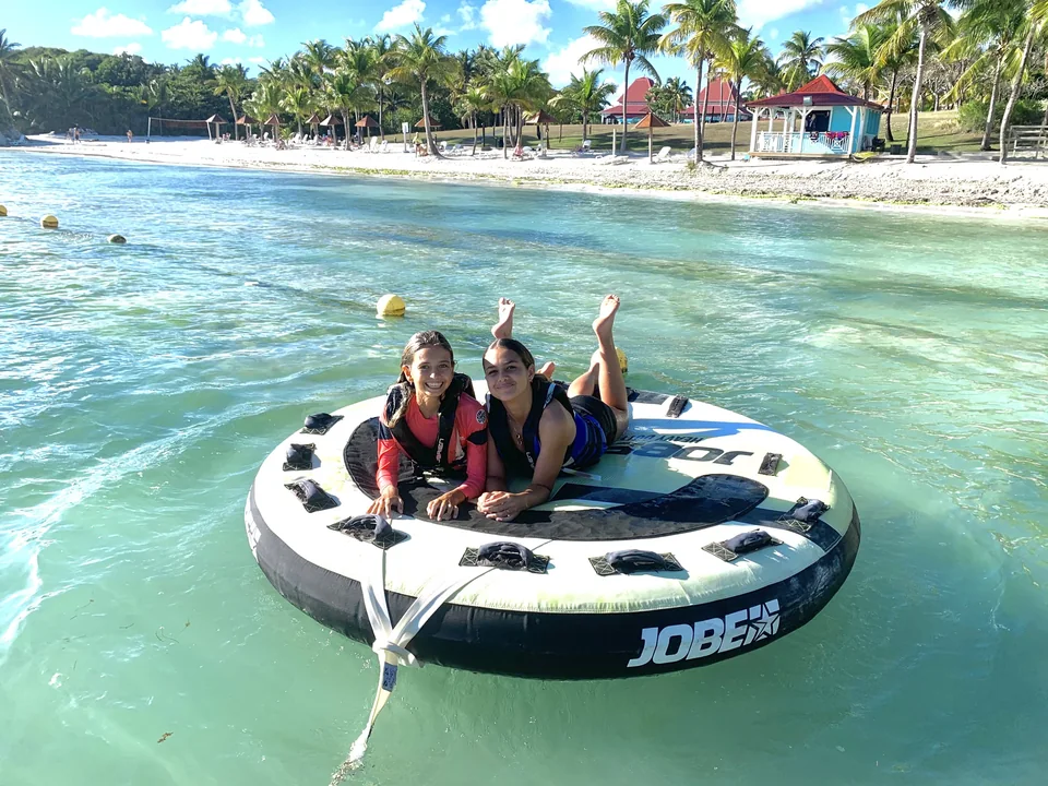 Deux jeunes filles sur une bouée tractée Atmosphère, sur l'eau turquoise devant une plage de cocotiers en Guadeloupe