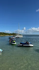 Jet skis alignés dans les eaux peu profondes, participants debout dans l’eau, voilier au mouillage