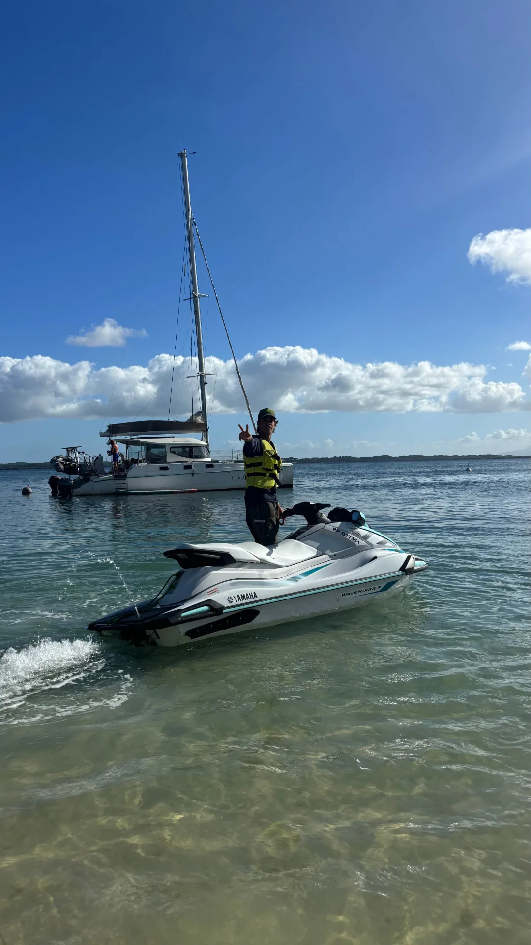 Moniteur en gilet jaune sur un jet ski, voilier au mouillage derrière lui